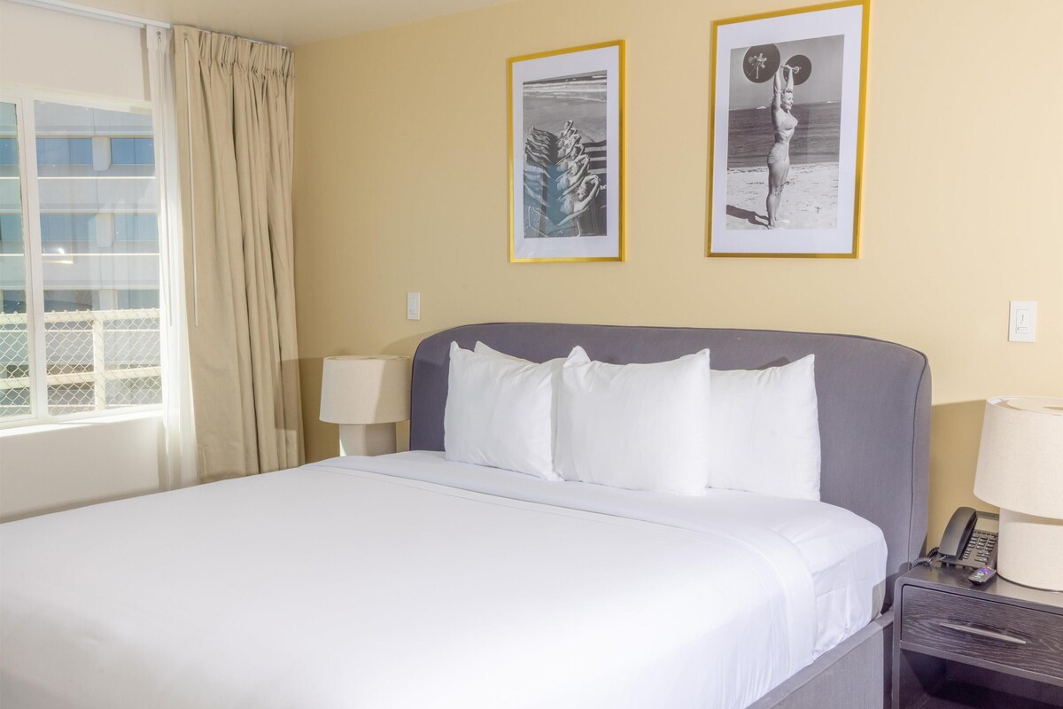A hotel room with a bed, two framed pictures on the wall, a window with curtains, and a bedside table with a lamp and phone.