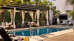 A pool area with lounge chairs, a pergola with white curtains, and a white building in the background. - The Eden (Santa Monica)