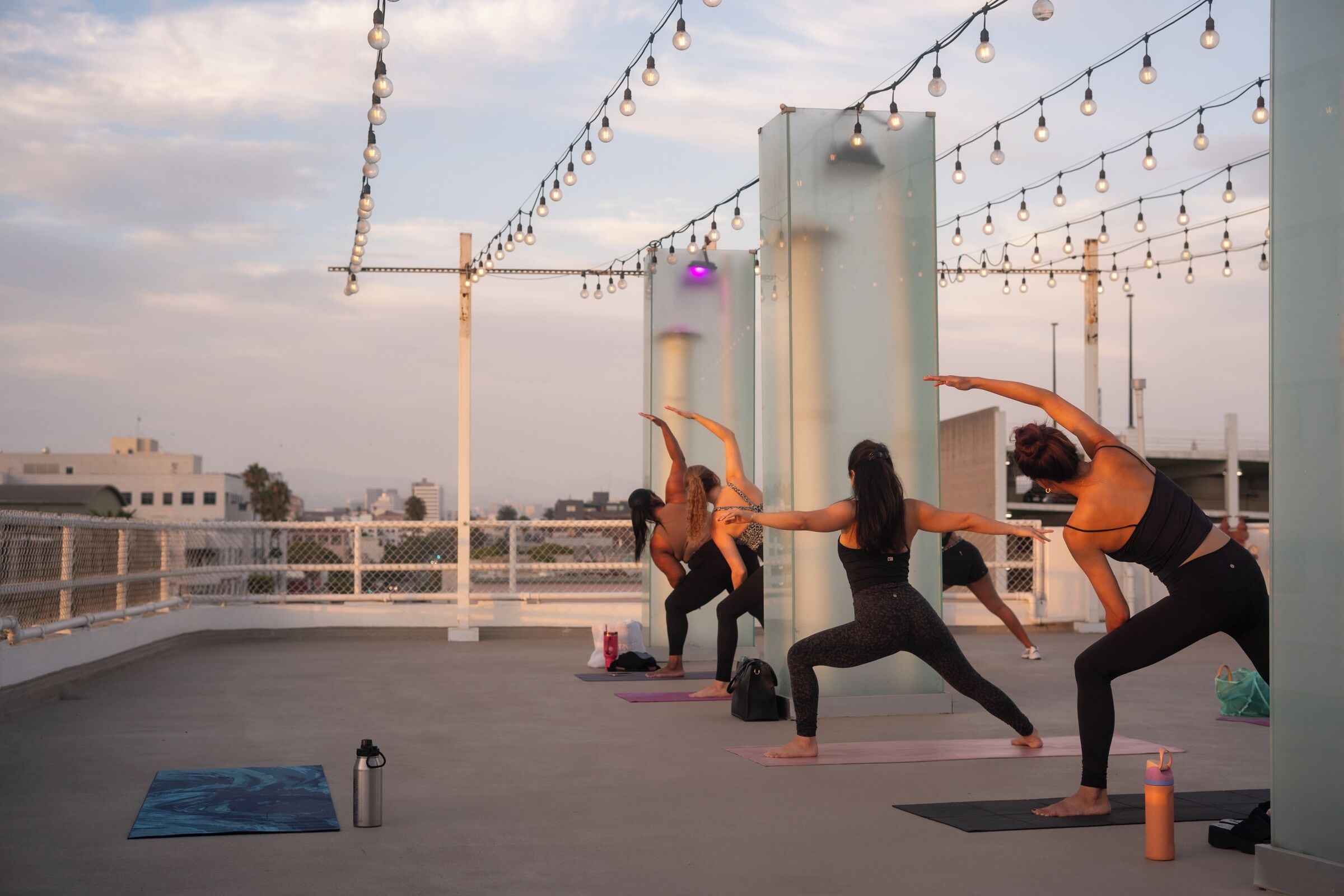 A rooftop yoga class with participants on mats, string lights overhead, and a cityscape in the background.