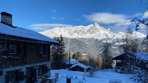 Un village enneigé, avec ses maisons traditionnelles en bois, un bâtiment en pierre doté d’une cheminée, et une chaîne de montagnes en arrière-plan.