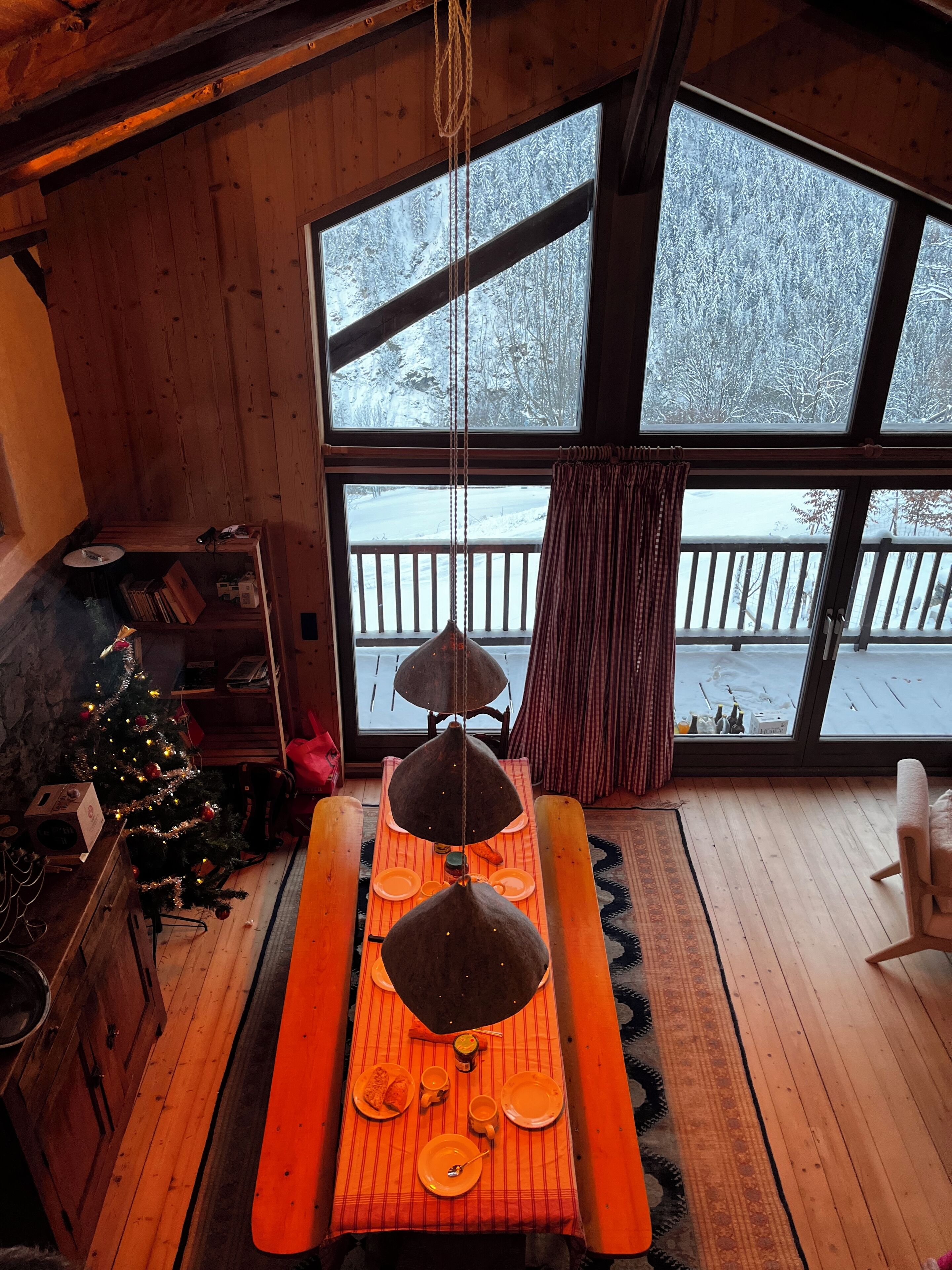 A cozy room with a wooden interior, a Christmas tree, a dining table set with plates and cups, and a large window overlooking a snowy deck.