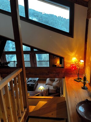 Interior acolhedor de uma cabana com quarto no mezanino, área de jantar e vista para uma floresta nevada através de grandes janelas.