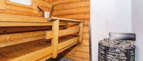 A sauna room with wooden benches, a metal bucket, and a stone-filled container.