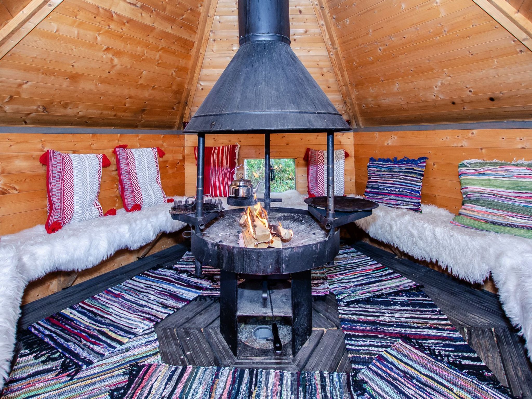 A cozy cabin interior with a central wood-burning stove, plush seating with patterned cushions, and a wooden ceiling.
