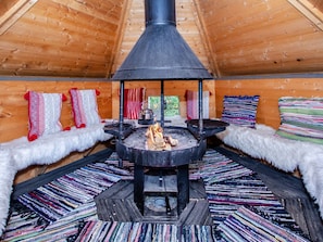 A cozy cabin interior with a central wood-burning stove, plush seating with patterned cushions, and a wooden ceiling.