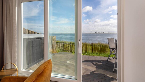 Una habitación con puerta corrediza de vidrio que da a una terraza con vista al mar y aerogeneradores a lo lejos.