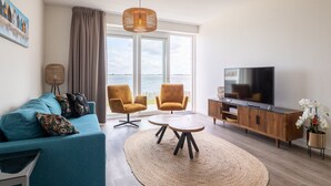 Una sala de estar moderna con un sofá, dos sillones, una mesa de centro, un televisor y un amplio ventanal con vistas al mar.
