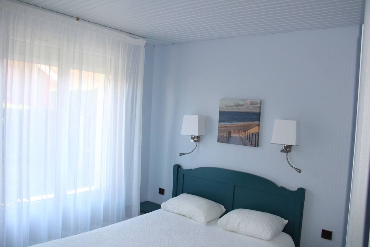 Une chambre d’hôtel avec un lit, des rideaux blancs, deux lampes fixées au mur et un tableau accroché au mur.