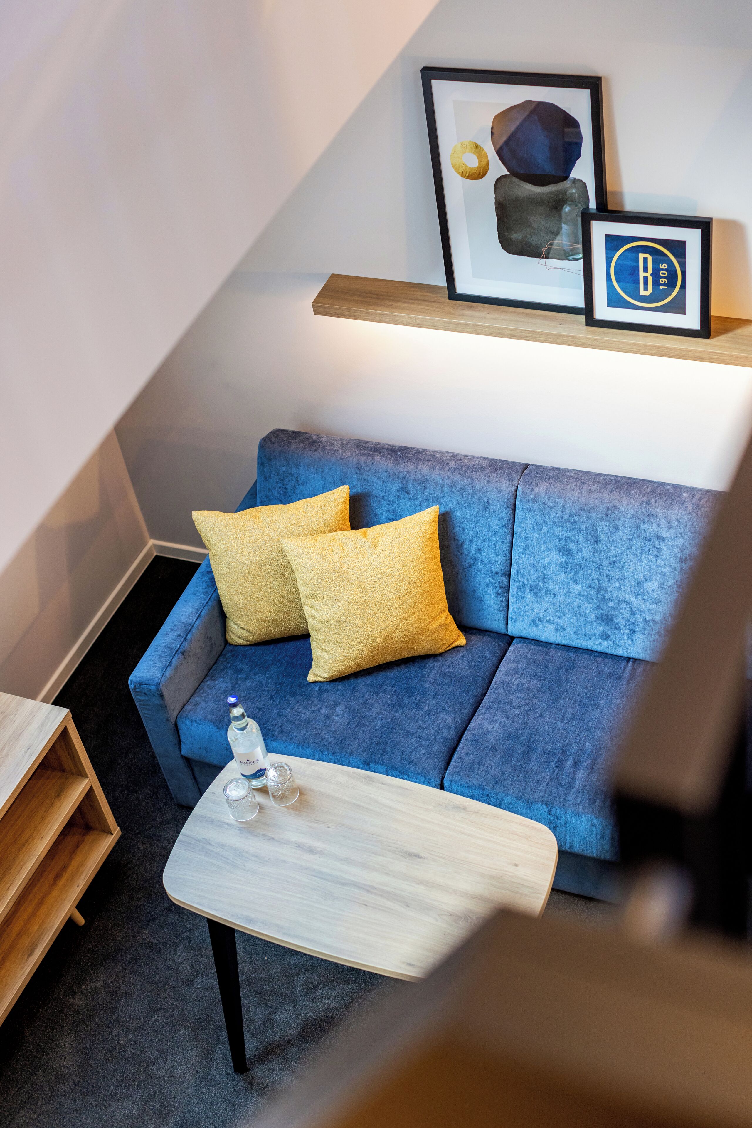 Ein blaues Sofa mit gelben Kissen, ein kleiner Holztisch und ein gerahmtes abstraktes Kunstwerk auf einem Regal darüber.