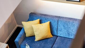 Ein blaues Sofa mit gelben Kissen, ein kleiner Holztisch und ein gerahmtes abstraktes Kunstwerk auf einem Regal darüber.