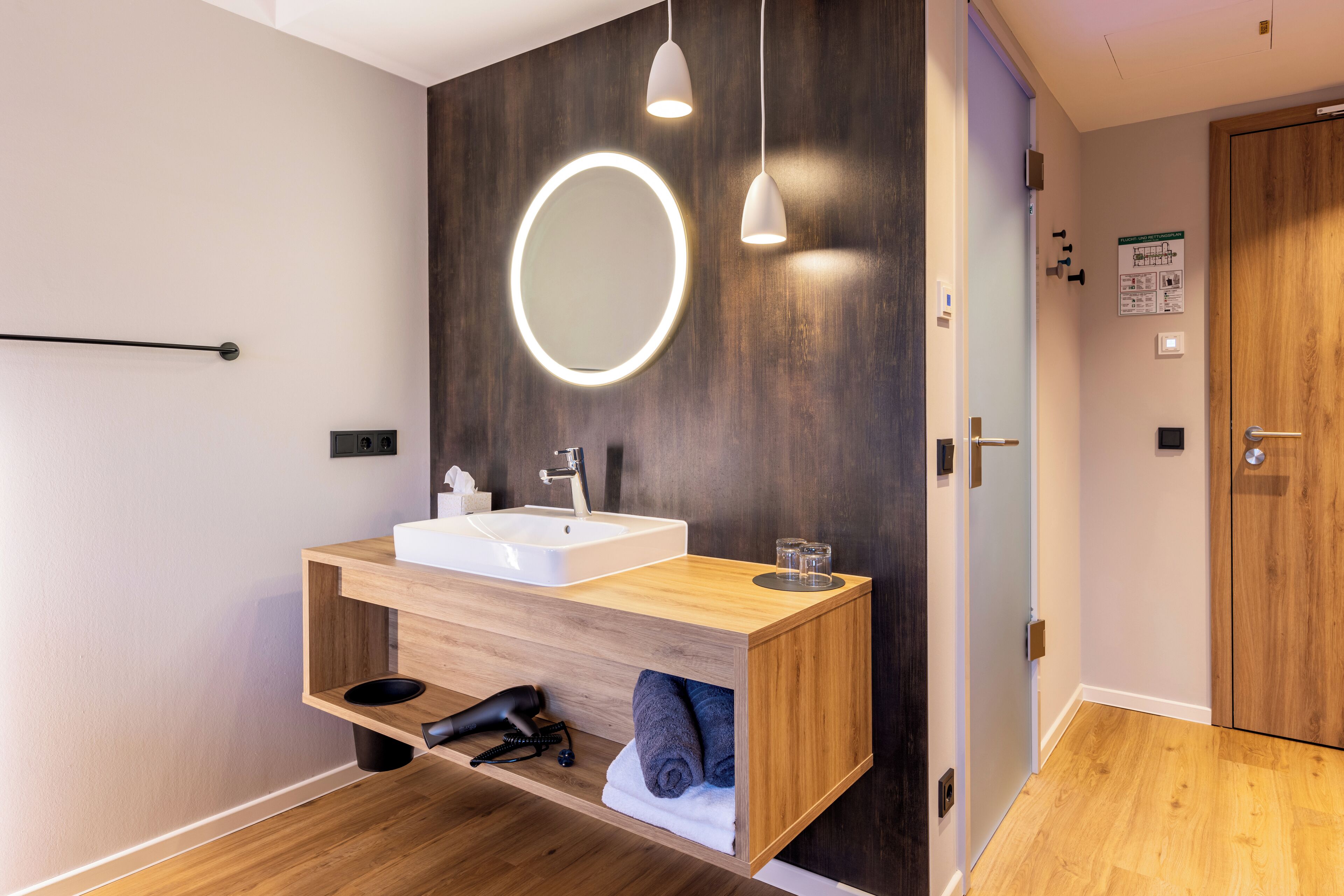 Un baño moderno con tocador de madera, espejo redondo e iluminación colgante.