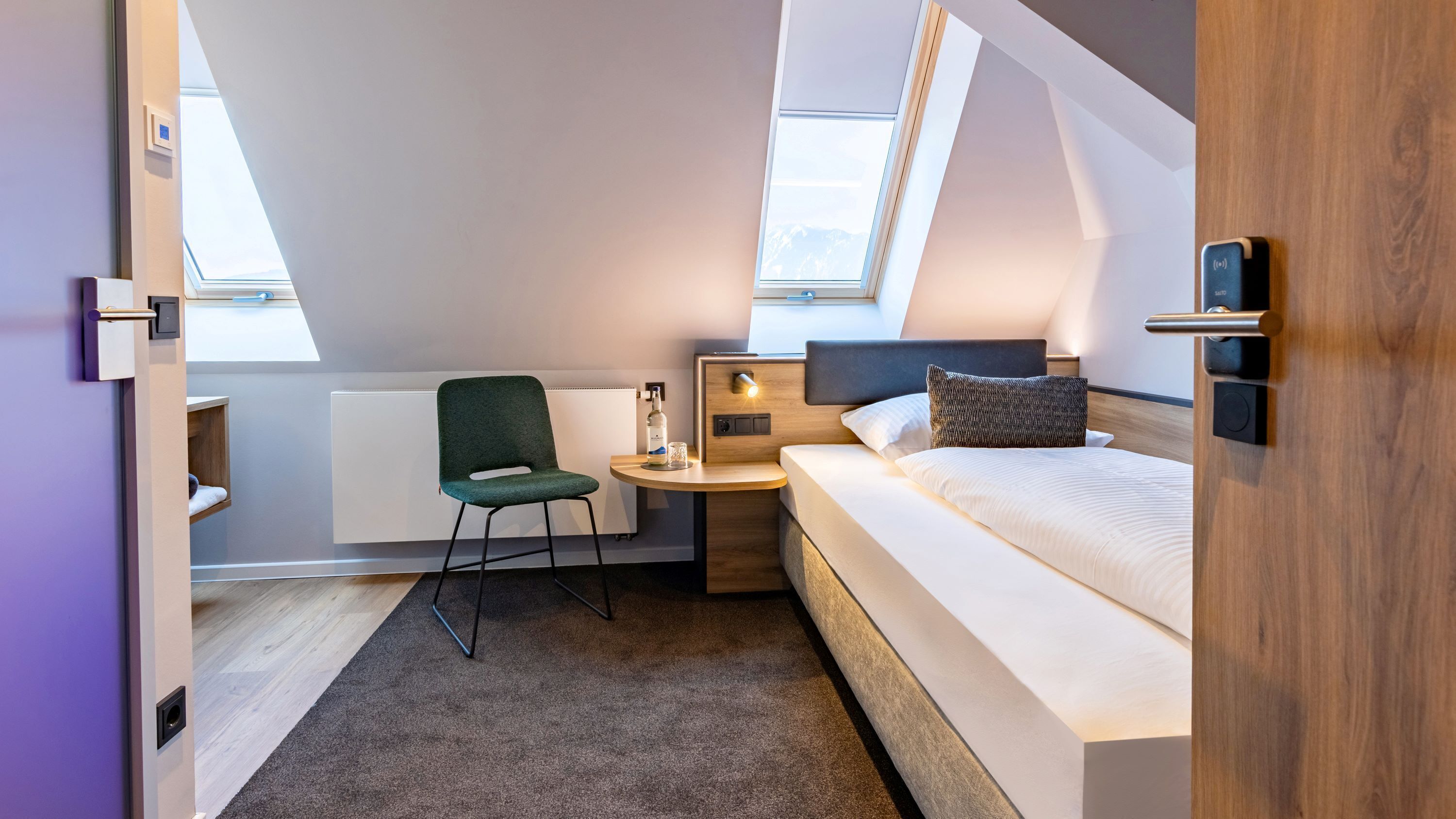A compact hotel room with a bed, a small desk, a chair, and a window.