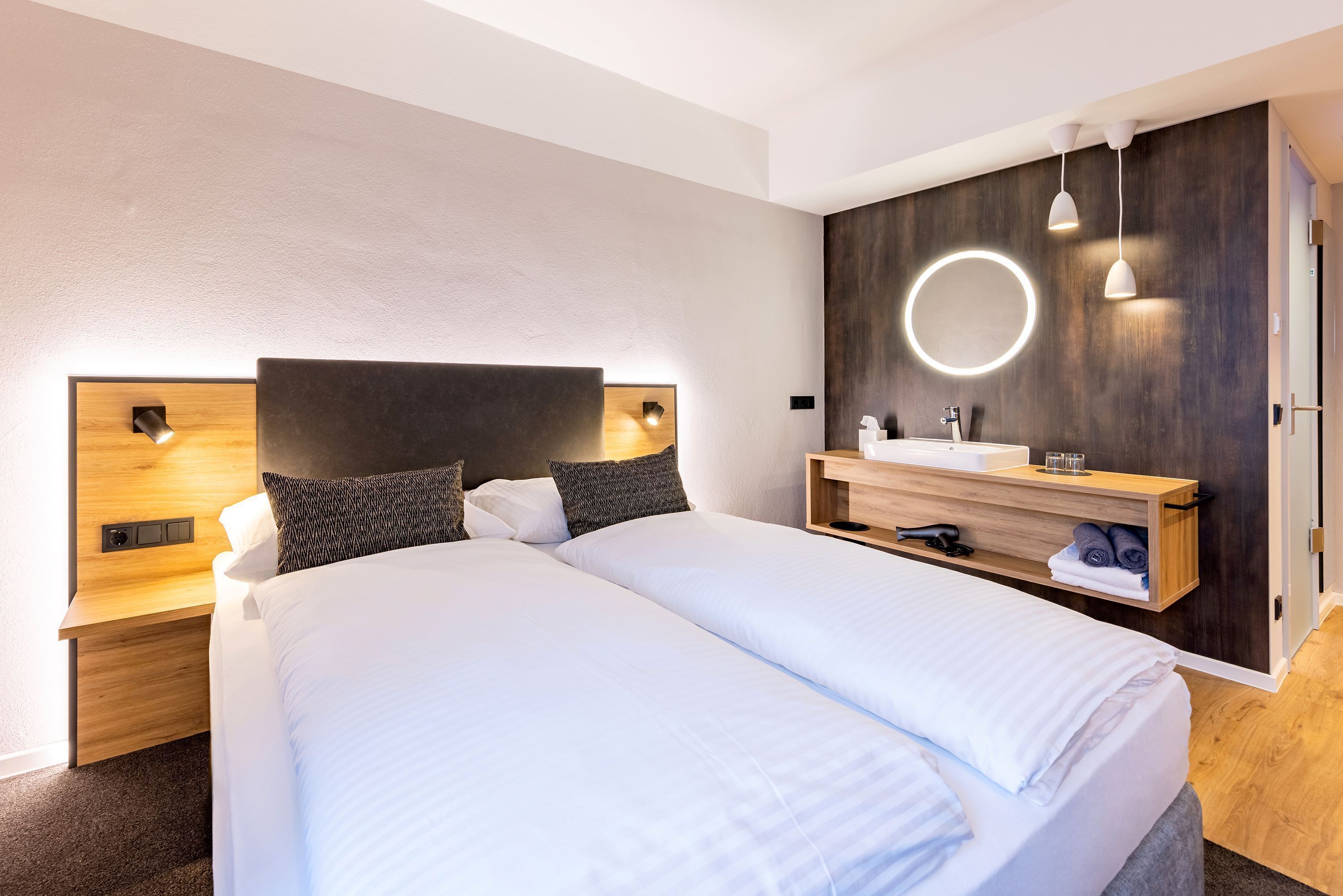A modern hotel room with a large bed, a wooden headboard, a built-in vanity with a mirror, and a small shelf with towels.