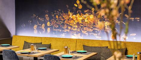 Interior de un restaurante con una larga mesa de madera, asientos acolchados y una decoración de monedas en la pared.