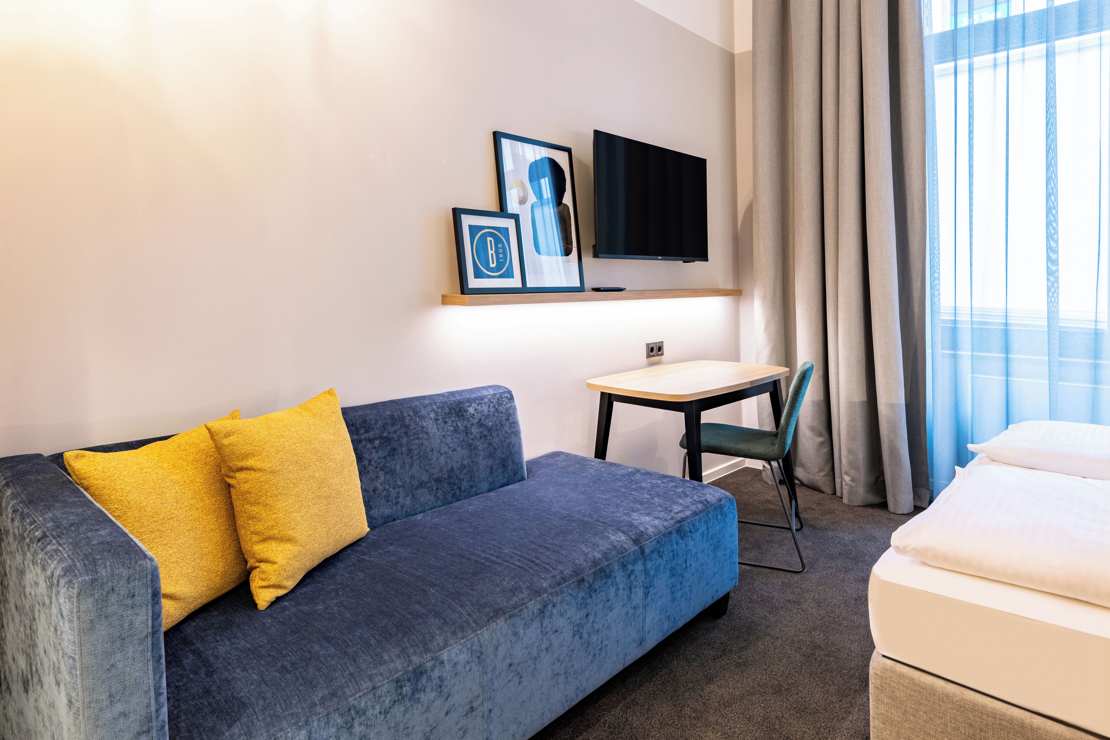 A hotel room with a blue sofa, a small table, a TV mounted on the wall, and a bed.