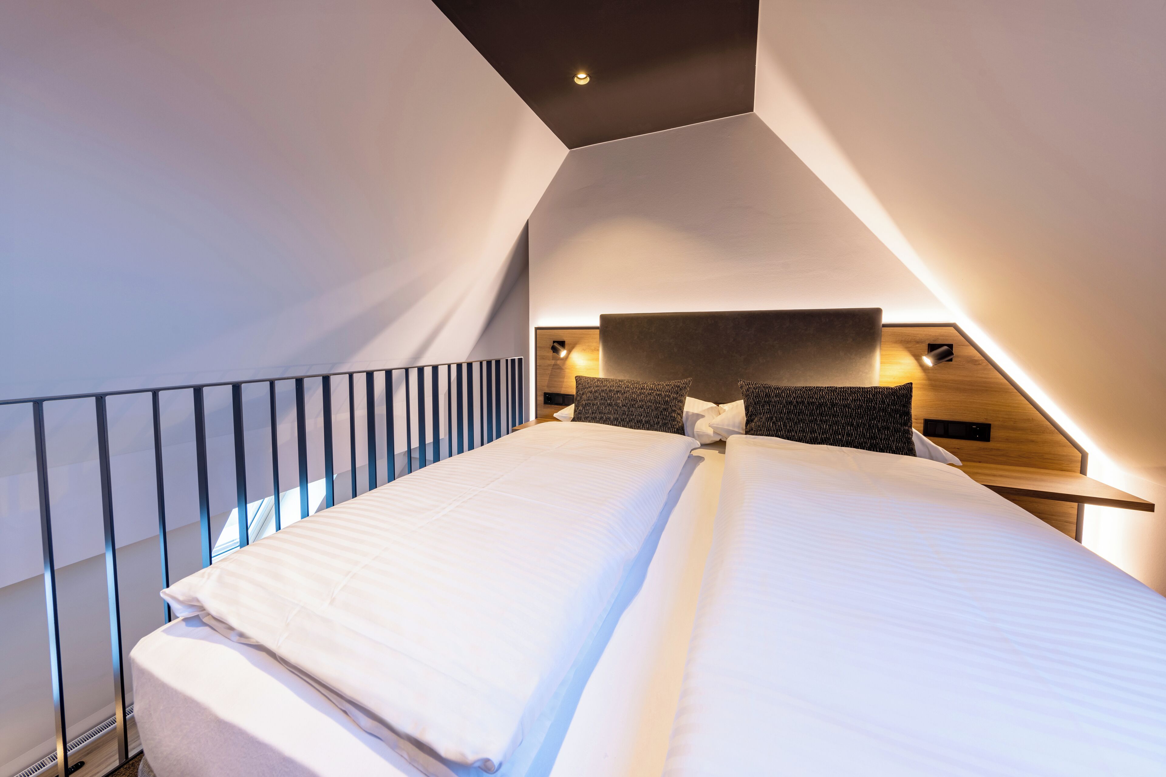 Una habitación de hotel moderna con techo inclinado, una cama grande con ropa blanca, dos mesitas de noche con lámparas y vistas al exterior.