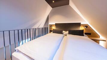 Una habitación de hotel moderna con techo inclinado, una cama grande con ropa blanca, dos mesitas de noche con lámparas y vistas al exterior.