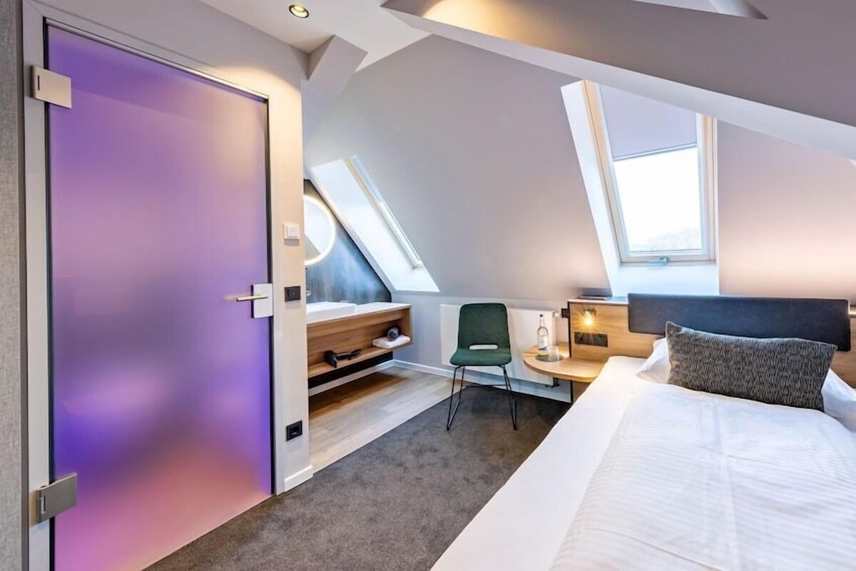 A modern hotel room with a bed, a desk, a chair, and a purple-framed door.