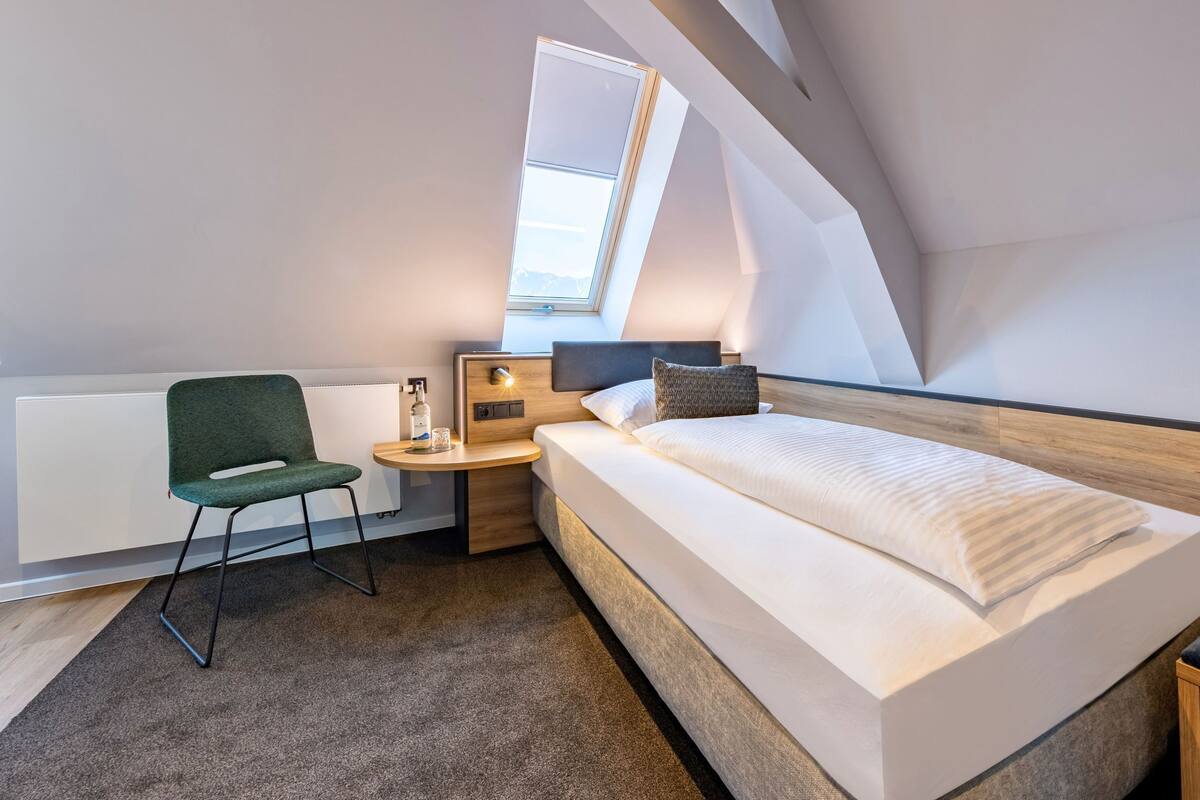 A modern, compact hotel room with a skylight, a single bed, a small desk, a chair, and a bedside table.