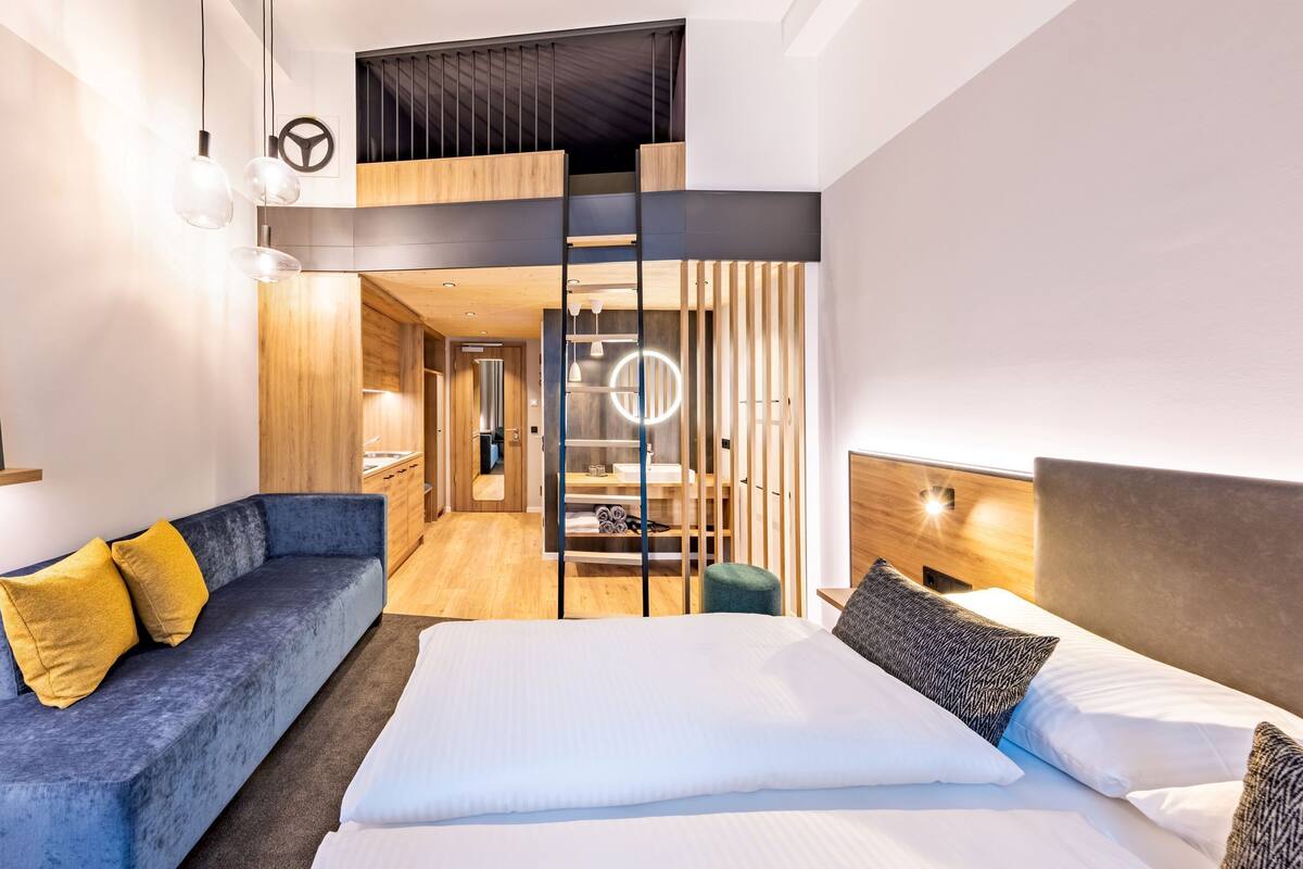 A modern hotel room with a bed, a sofa, a mirror, and a bathroom visible through a partition.