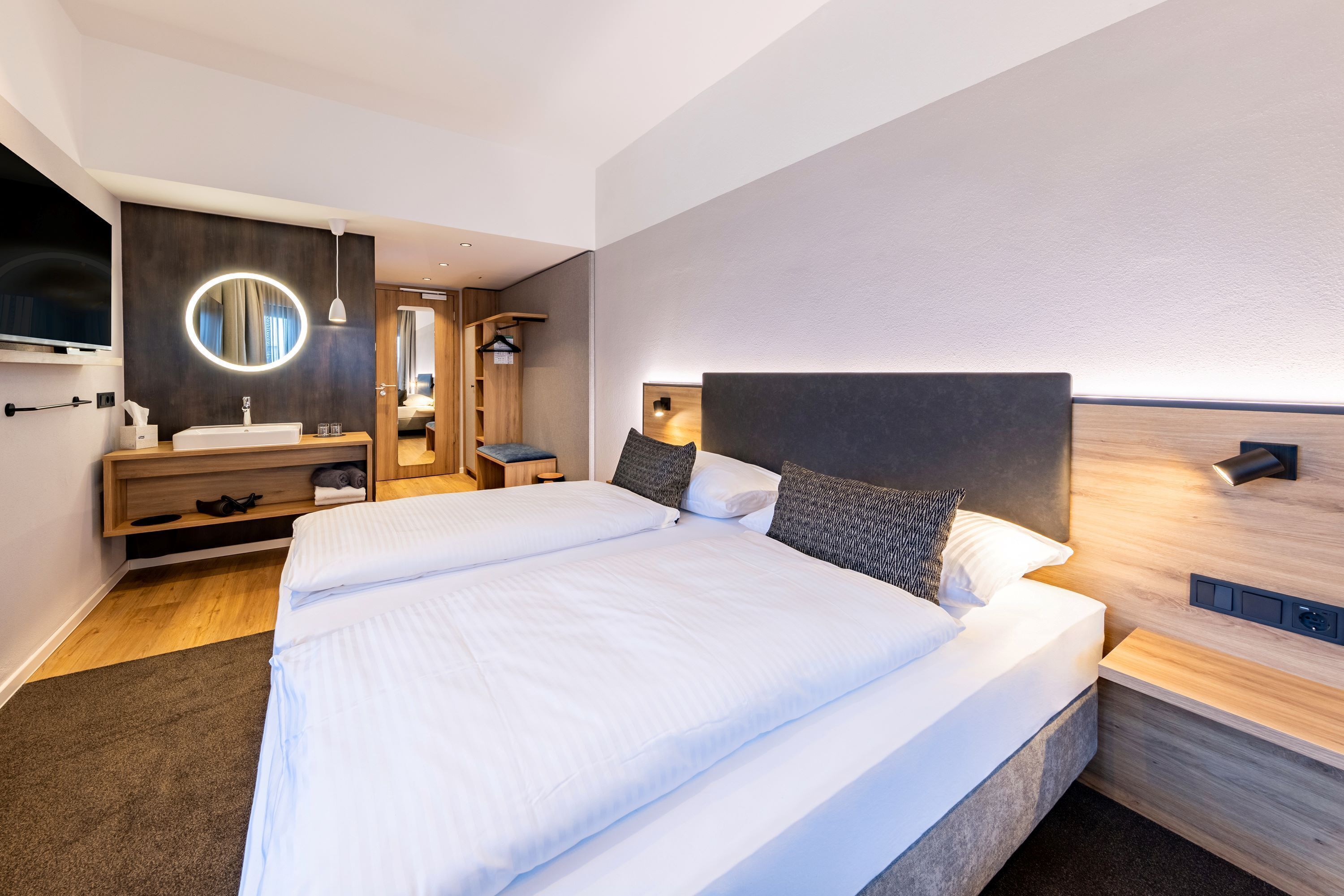 Ein modernes Hotelzimmer mit einem großen Bett, einem runden Spiegel, einer Holzkommode und einem an der Wand montierten Fernseher.