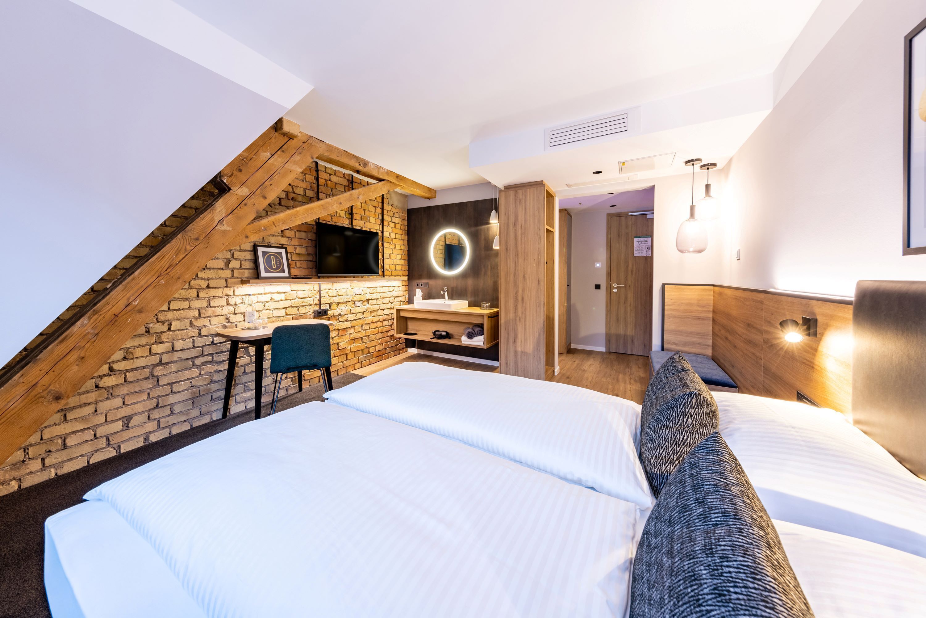 Ein modernes Hotelzimmer mit einer erhöhten Schlafkoje, einem kleinen Schreibtisch und einem Badezimmer mit Spiegel und Waschbecken.