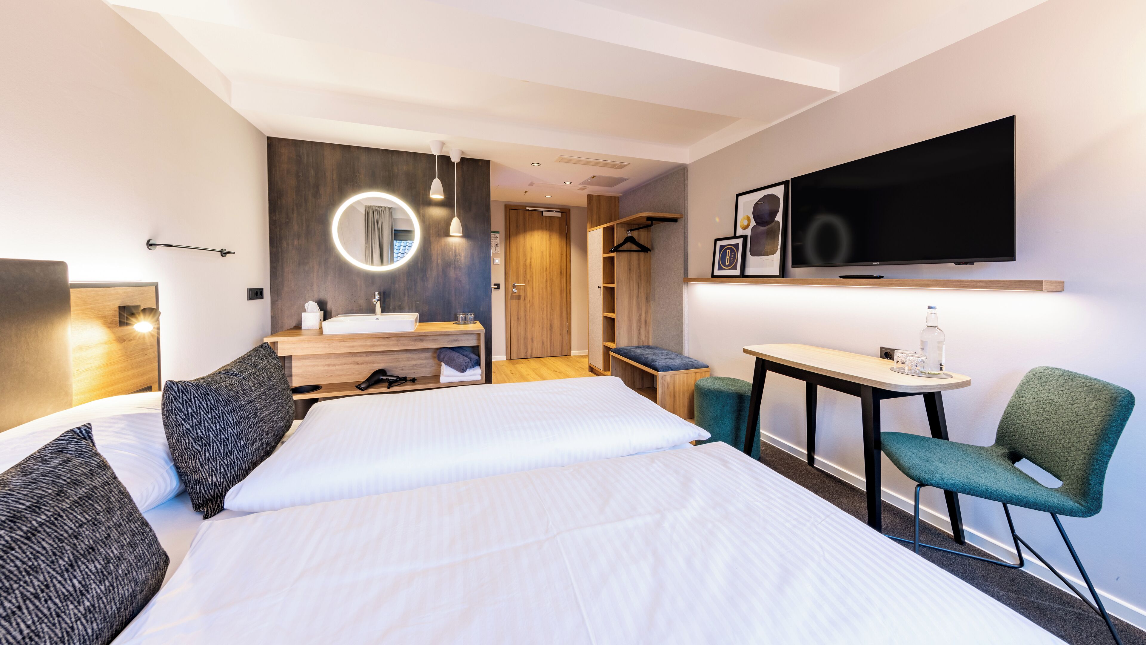 A modern hotel room with a large bed, a desk with a chair, a TV, a mirror, and a wardrobe.