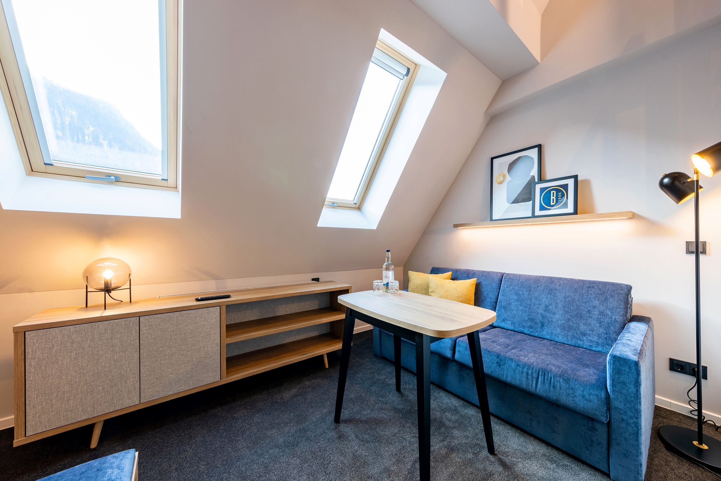 A modern loft bedroom with skylights, a blue sofa, a small table, and a desk with a lamp.