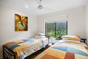 A room with two single beds, a ceiling fan, a large window with a view of greenery, and a painting on the wall.