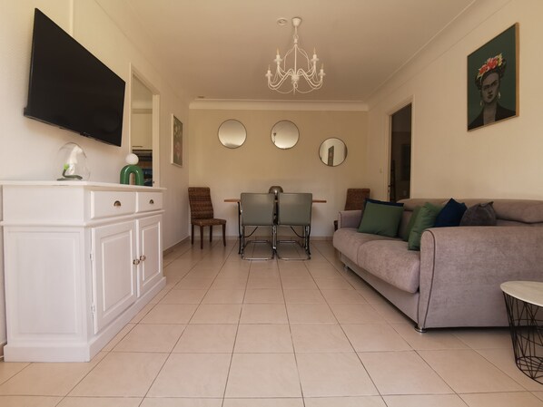 A living room with a sofa, a dining table with chairs, a TV mounted on the wall, and a chandelier.