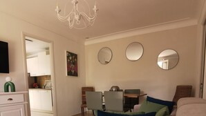 Ett vardagsrum med en beige soffa, ett matbord med stolar, en kristallkrona och ett kök i bakgrunden.