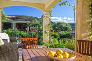 Un patio techado con mobiliario de mimbre, una mesa de madera con un cuenco de fruta, y una vista de una casa con techo de tejas y vegetación al fondo.