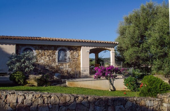 Un edificio de piedra con techo de tejas, un muro de piedra y un jardín con flores moradas.