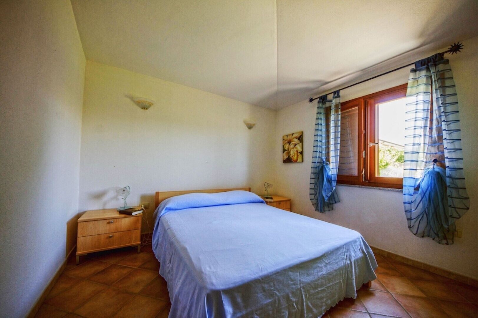 Een slaapkamer met een enkel bed, houten nachtkastjes, een wandlamp, een raam met gordijnen en een klein schilderij.