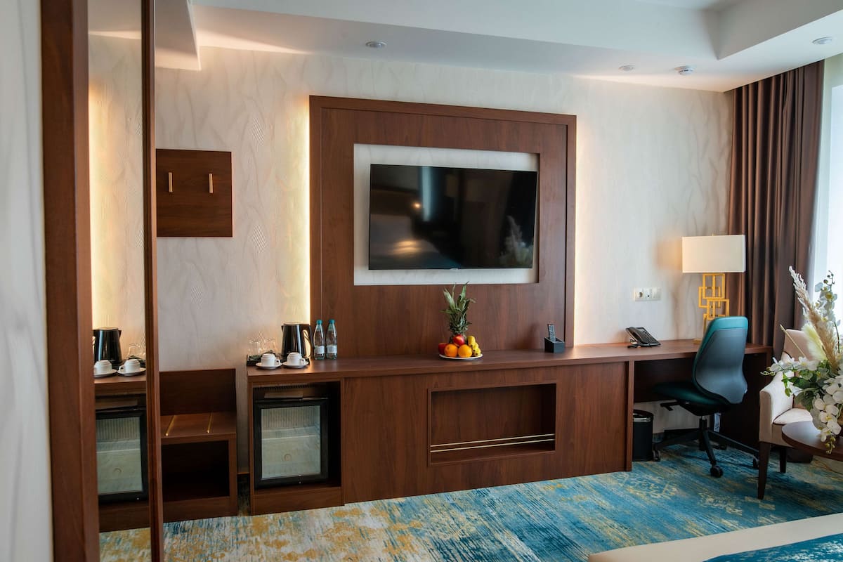 A hotel room with a wooden desk, a flat-screen TV, a chair, a small shelf with a pineapple and oranges, a lamp, and a decorative plant.