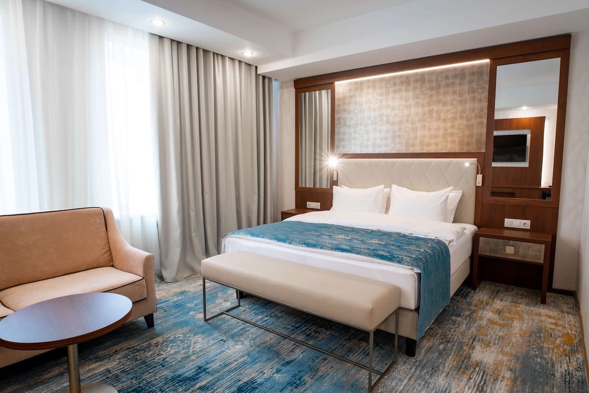 A hotel room with a bed, a bench, a chair, a bedside table, and a large mirror.