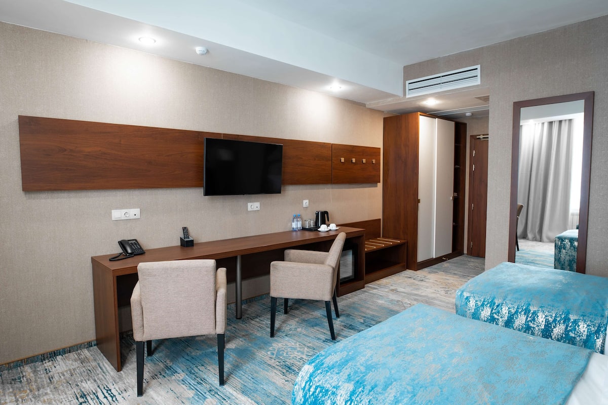 A hotel room with a desk, two chairs, a TV, a bed with blue bedding, and a wardrobe.