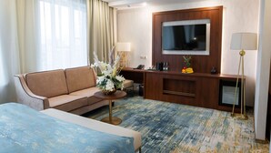 A hotel room with a sofa, a small round table with a vase, a flat-screen TV mounted on the wall, a desk with a lamp, and a bed with a blue cover. - Best Western Plus Oskemen (Oskemen)