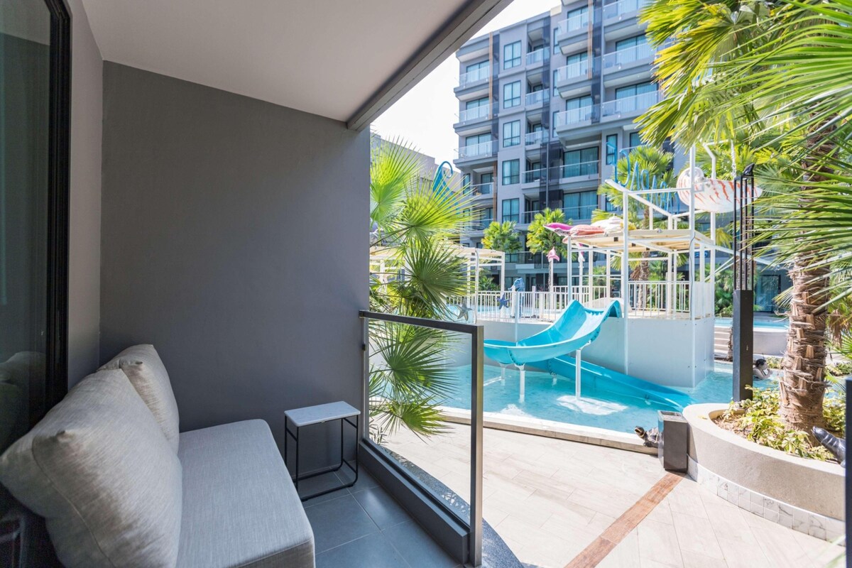 A balcony with a sofa, a small table, and a view of a swimming pool with a slide.
