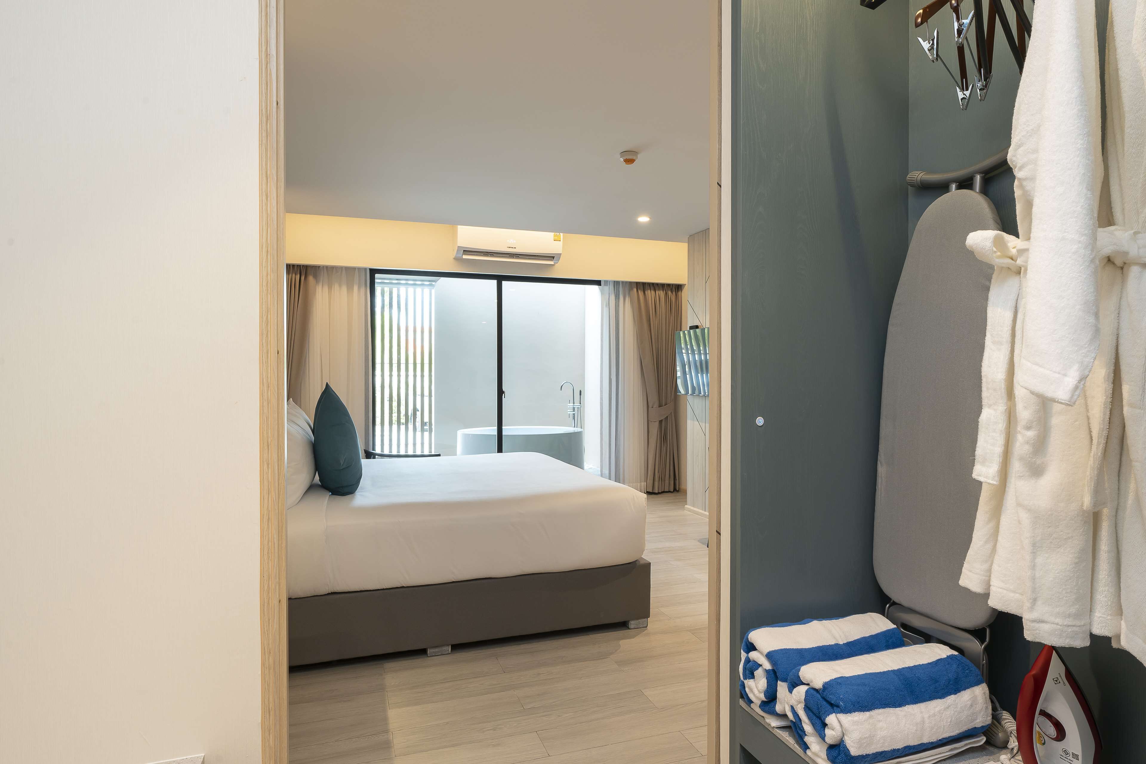 Una habitación de hotel con cama, armario, tabla de planchar y ventana con cortinas.