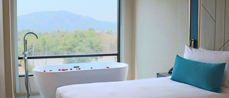 A bedroom with a large bed, a bathtub filled with rose petals, and a view of mountains through a large window.