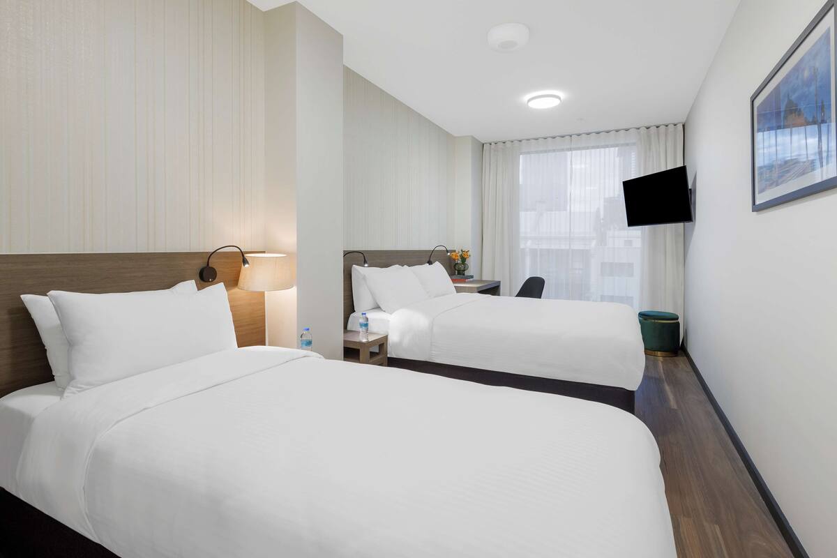 Een hotelkamer met twee bedden, een bureau, een stoel, een aan de muur bevestigde televisie en een raam met gordijnen.