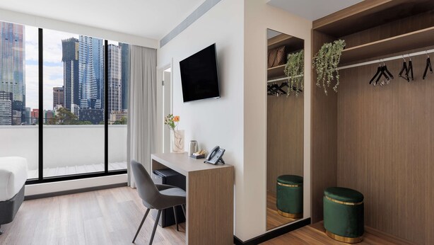 Habitación de hotel moderna con un ventanal grande, un escritorio, una silla, un televisor, un armario y un taburete verde pequeño.