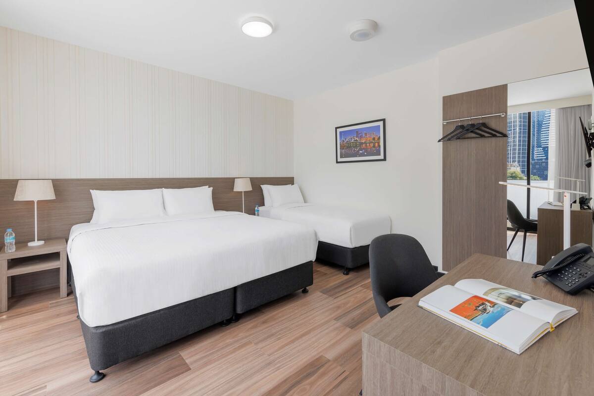 Een hotelkamer met een groot bed, twee kleinere bedden, een bureau met telefoon, een stoel, een lamp en een ingelijst schilderij aan de muur.