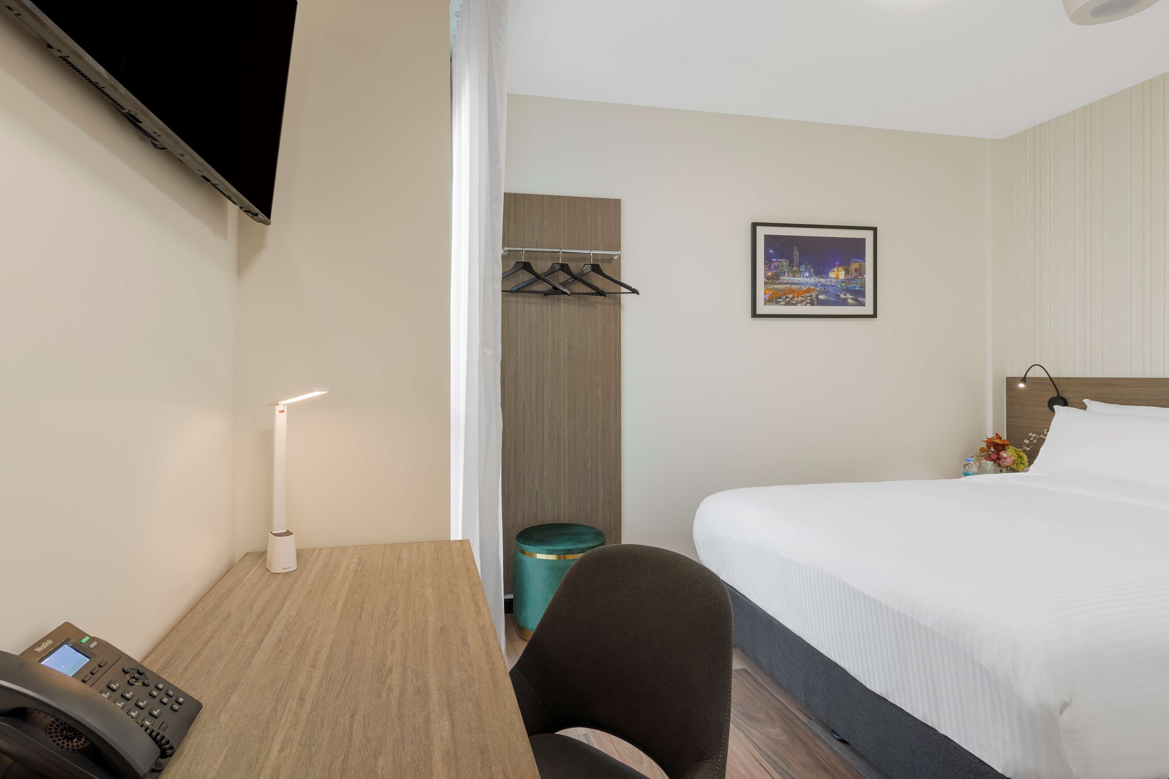 A hotel room with a bed, a desk with a telephone, a lamp, a picture on the wall, and a coat rack.