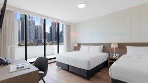 Habitación de hotel con dos camas, un escritorio y una silla. Cuenta con un ventanal grande con vistas a la ciudad.