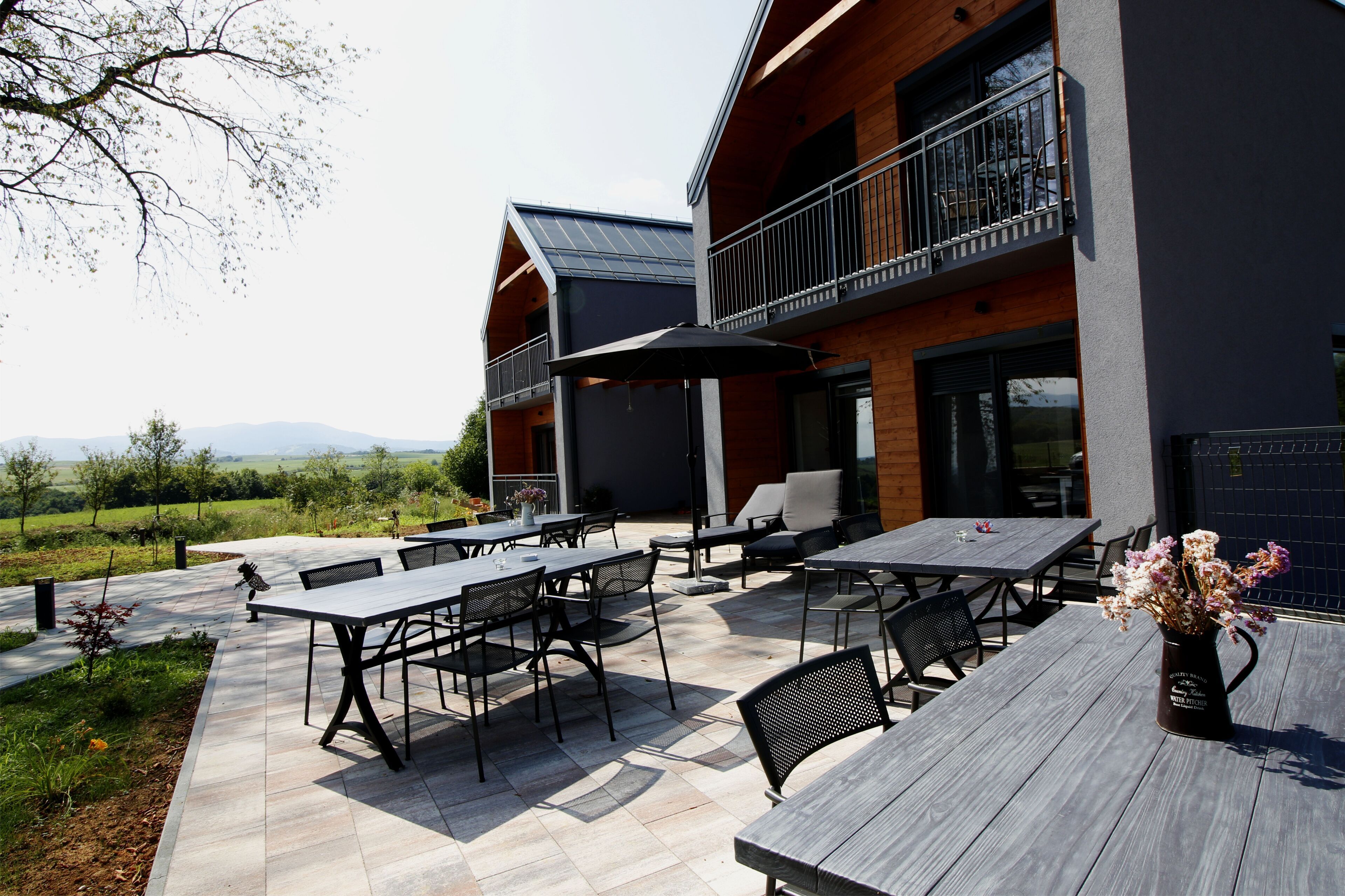 An outdoor patio area with multiple tables and chairs, a wooden fence, and a view of the countryside.