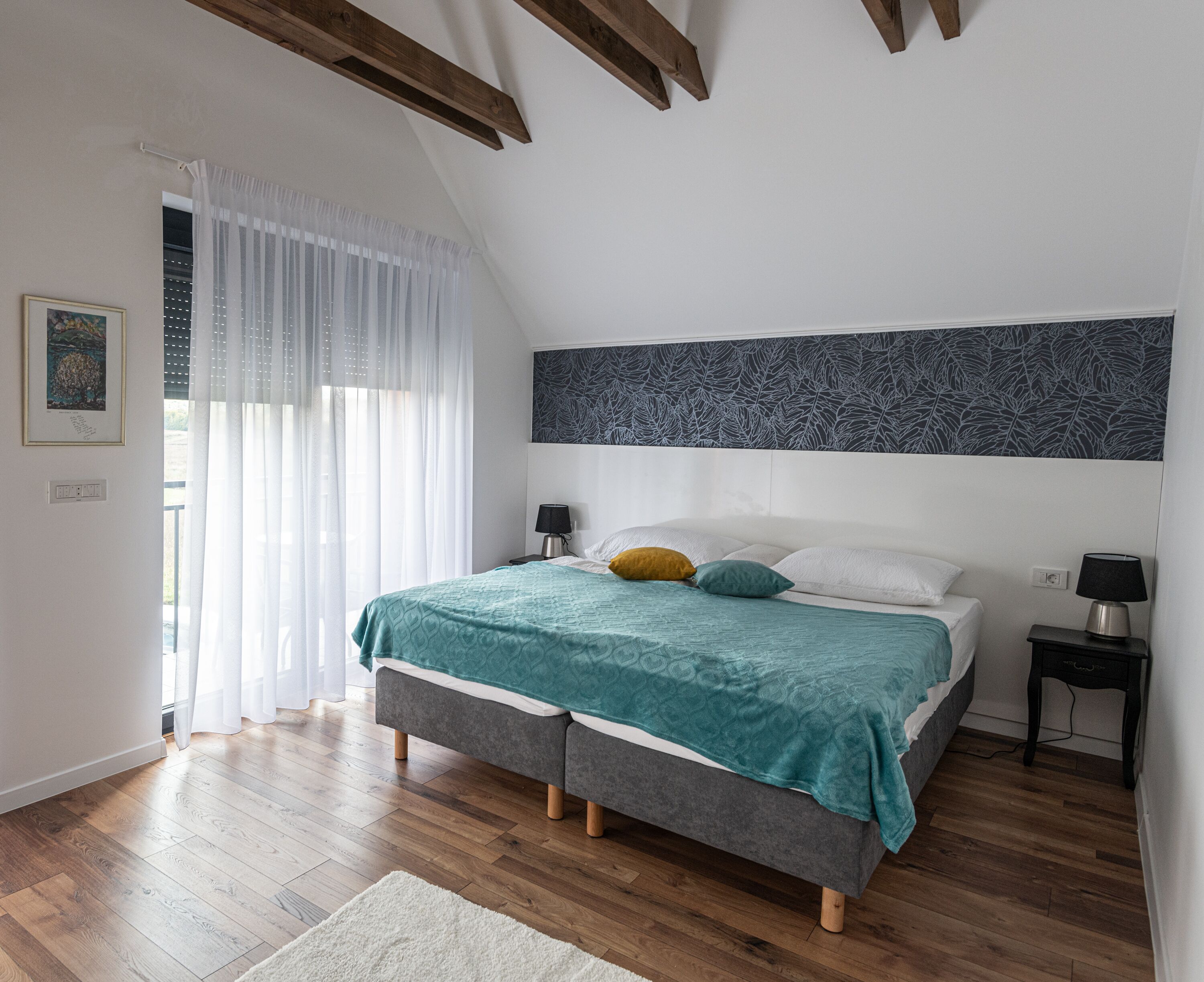 A bedroom with a bed, two bedside tables, a window with sheer curtains, and a ceiling with exposed beams.