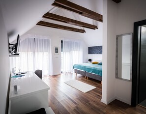 Un dormitorio moderno con techo inclinado, vigas de madera, cama con colcha color turquesa, un escritorio blanco con computadora y un espejo grande.
