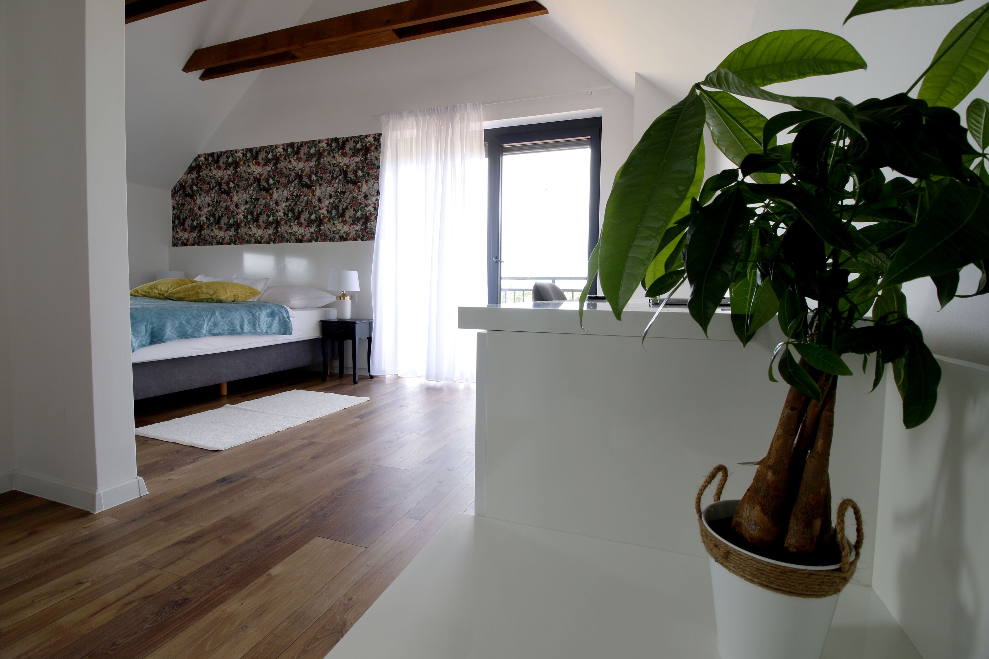Un dormitorio con cama, mesita de noche, lámpara, ventana con cortinas y una planta grande en maceta.